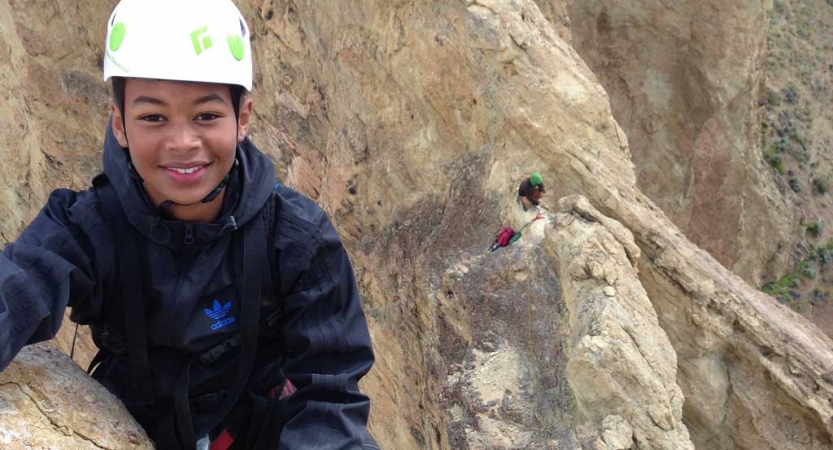 A young person wearing safety gear smiles at the camera amongst a rocky landscape.